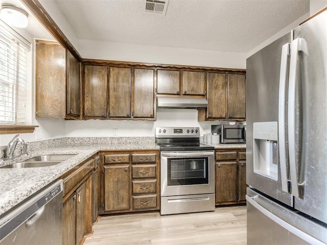kitchen with appliances with stainless steel finishes, a sink, under cabinet range hood, light wood-style flooring, and a textured ceiling
