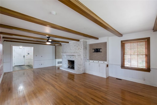 1301 W C Ave, Elk City, OK 73644 - unfurnished living room featuring a ceiling fan, wood finished floors, beamed ceiling, a brick fireplace, and a wainscoted wall