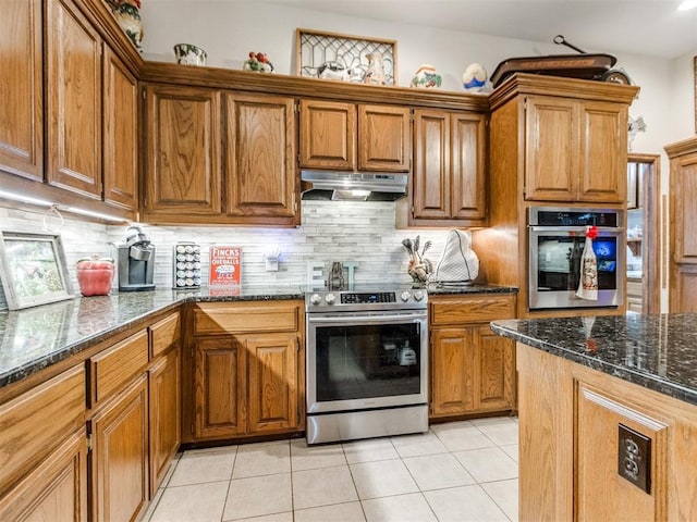19609 Filly Dr, Edmond, OK 73003 - kitchen featuring appliances with stainless steel finishes, under cabinet range hood, brown cabinetry, light tile patterned floors, and dark stone counters