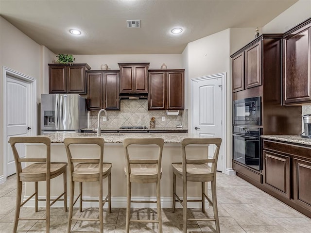 2500 Cordgrass Ln, Edmond, OK 73013 - kitchen featuring black appliances, under cabinet range hood, a kitchen island with sink, backsplash, and dark brown cabinets