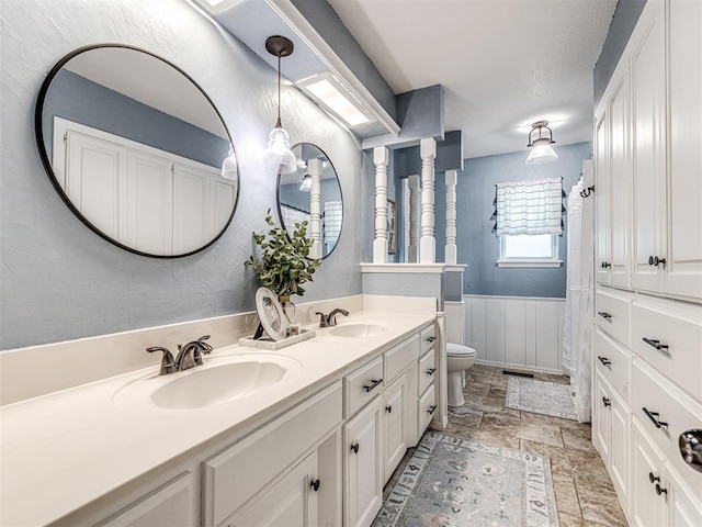 2009 Bella Vista Dr, Edmond, OK 73013 - full bath featuring toilet, double vanity, a wainscoted wall, and stone finish flooring