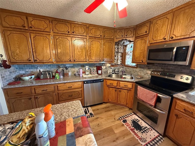 kitchen with appliances with stainless steel finishes, a sink, brown cabinetry, a textured ceiling, and light wood-type flooring