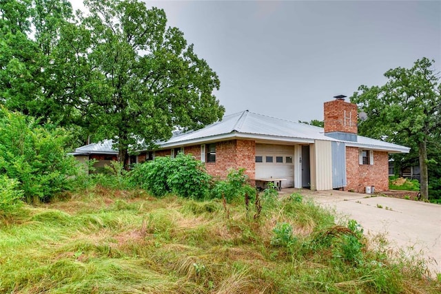 view of property exterior with an attached garage, a chimney, driveway, metal roof, and brick siding