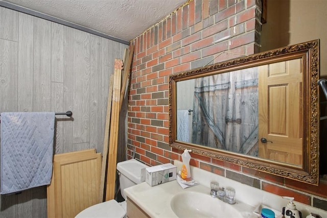 1001 N Pine St, Midwest City, OK 73130 - bathroom with a textured ceiling, toilet, vanity, brick wall, and wooden walls