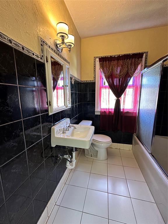 2001 NW 18th St, Oklahoma City, OK 73106 - full bath with a textured ceiling, tile patterned flooring, tile walls, a wainscoted wall, and bath / shower combo with glass door