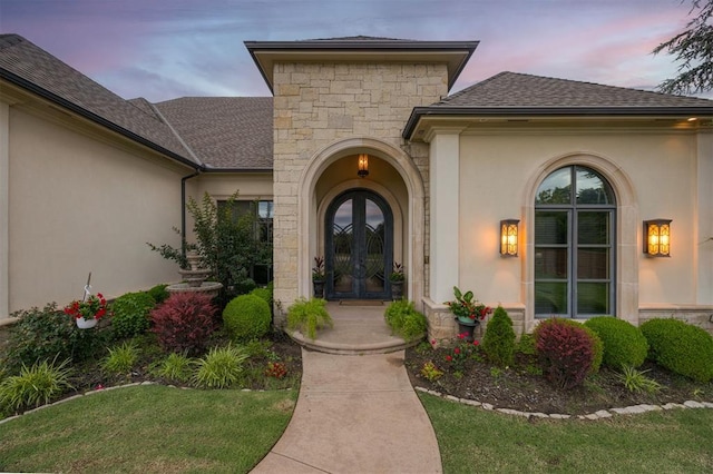 3617 Stagmoor Rd, Edmond, OK 73034 - exterior entry at dusk featuring french doors, stucco siding, a shingled roof, and stone siding