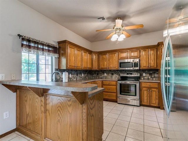 1204 Scrambler Ln, Edmond, OK 73025 - kitchen featuring stainless steel appliances, brown cabinets, a peninsula, light tile patterned floors, and decorative backsplash