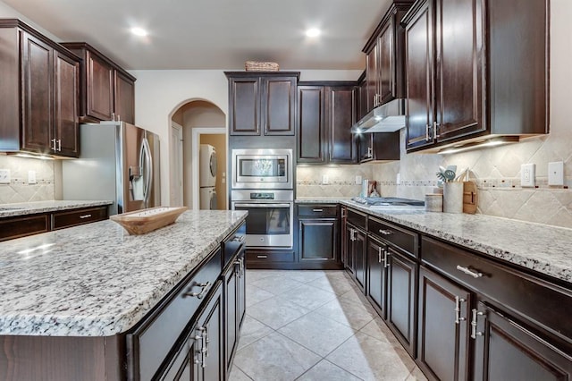 637 Branch Line Rd, Yukon, OK 73099 - kitchen with stainless steel appliances, stacked washer / dryer, under cabinet range hood, light stone countertops, and dark brown cabinets