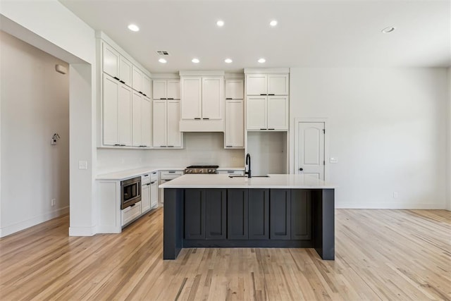 1690 Shaylee Ln, Choctaw, OK 73020 - kitchen featuring stainless steel microwave, a kitchen island with sink, white cabinetry, light wood-type flooring, and light countertops