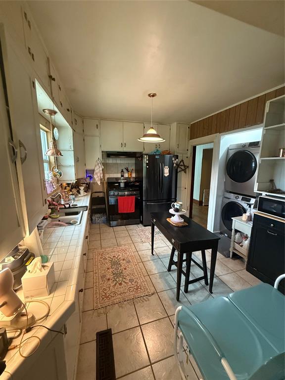 1522 S 9th St, Chickasha, OK 73018 - kitchen featuring tile countertops, stacked washer / drying machine, black appliances, light tile patterned flooring, and exhaust hood
