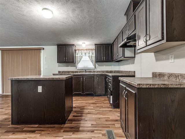 355370 E 970th Rd, Sparks, OK 74869 - kitchen with under cabinet range hood, a kitchen island, wood finished floors, a textured ceiling, and dark brown cabinets