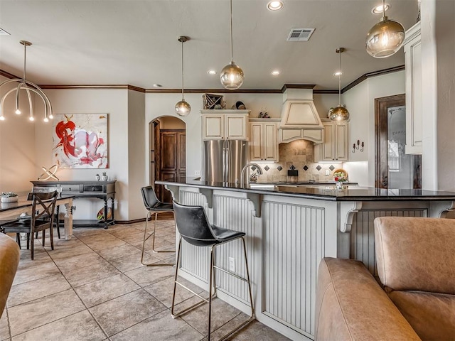 16208 Windrush Pl, Edmond, OK 73013 - kitchen with premium range hood, stainless steel fridge, cream cabinets, dark countertops, and backsplash