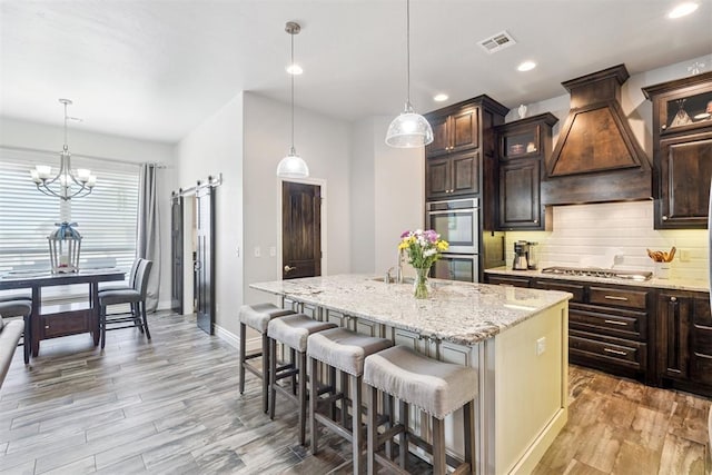 12505 Shady Gln, Choctaw, OK 73020 - kitchen featuring premium range hood, appliances with stainless steel finishes, an island with sink, light wood-type flooring, and dark brown cabinets
