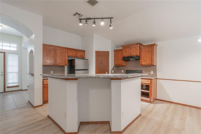 2412 NW 164th Ter, Edmond, OK 73013 - kitchen with freestanding refrigerator, under cabinet range hood, arched walkways, brown cabinetry, and decorative backsplash