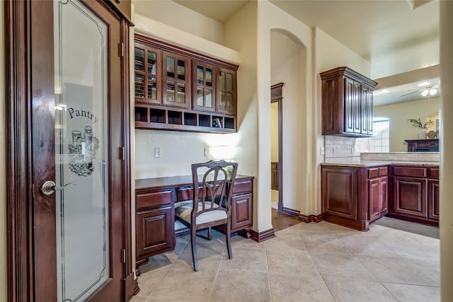 1704 NW 171st St, Edmond, OK 73012 - kitchen featuring built in desk, reddish brown cabinets, light tile patterned floors, a ceiling fan, and glass insert cabinets