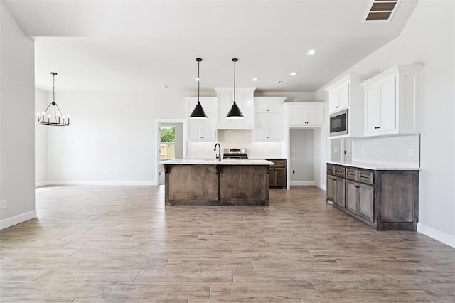10400 SW 51st St, Mustang, OK 73064 - kitchen featuring stainless steel microwave, light wood-style flooring, white cabinets, a kitchen island with sink, and a chandelier