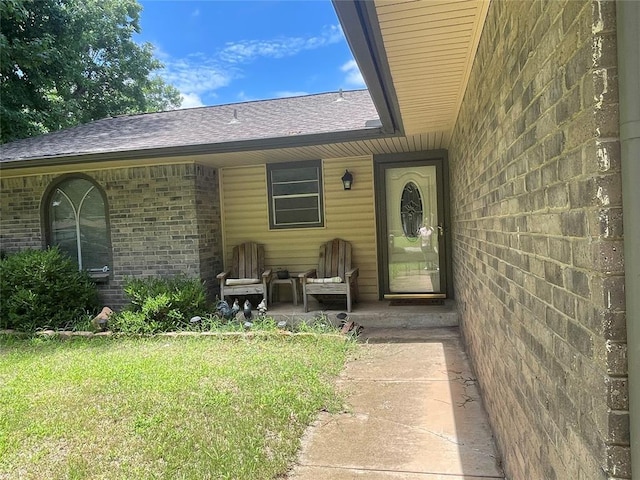 view of exterior entry featuring brick siding, a shingled roof, and a lawn