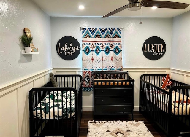 1717 S Webster Dr, Midwest City, OK 73130 - bedroom featuring a decorative wall, wood finished floors, a wainscoted wall, a crib, and a ceiling fan