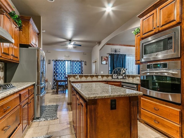 kitchen with appliances with stainless steel finishes, arched walkways, a kitchen island, tasteful backsplash, and brown cabinets