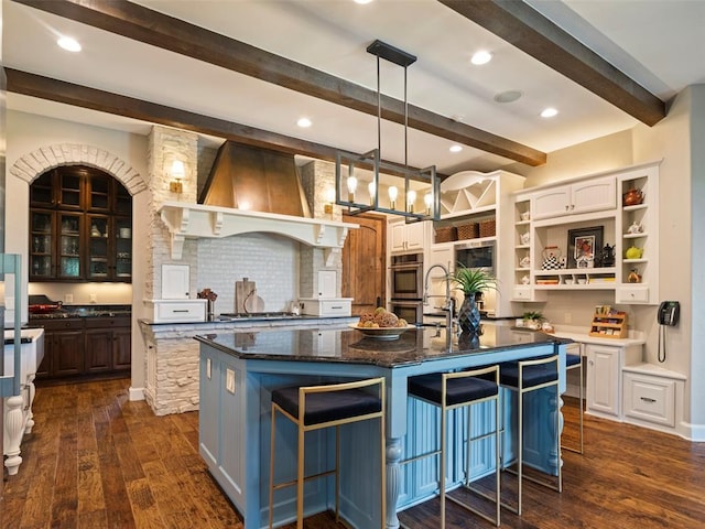 3325 Winding Creek Cir, Edmond, OK 73034 - kitchen featuring open shelves, beamed ceiling, custom range hood, a center island with sink, and dark wood finished floors