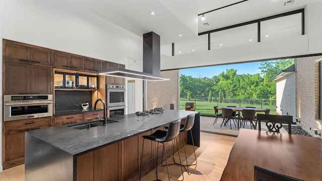 10105 SW 25th Ct, Yukon, OK 73099 - kitchen featuring a towering ceiling, stainless steel double oven, light wood-style flooring, an island with sink, and dark stone countertops