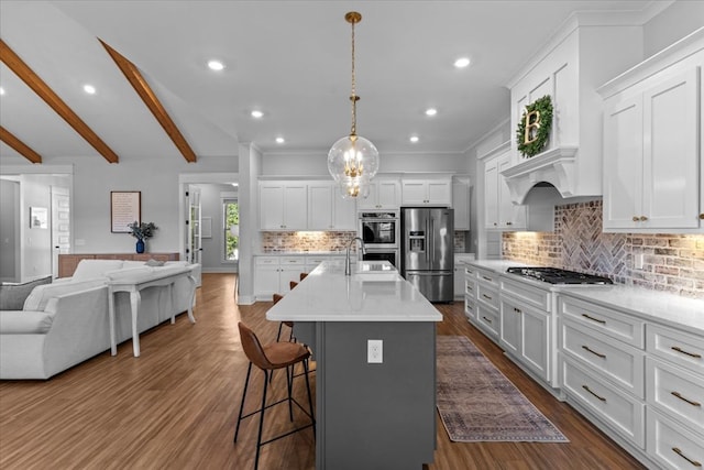 kitchen with white cabinetry, hanging light fixtures, beamed ceiling, an island with sink, and appliances with stainless steel finishes