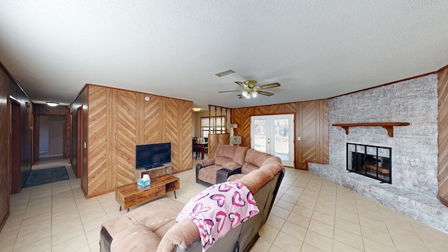 tiled living room with ceiling fan, french doors, a large fireplace, wood walls, and a textured ceiling