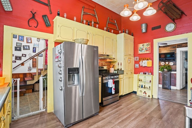 kitchen with light wood finished floors, a towering ceiling, appliances with stainless steel finishes, under cabinet range hood, and an inviting chandelier