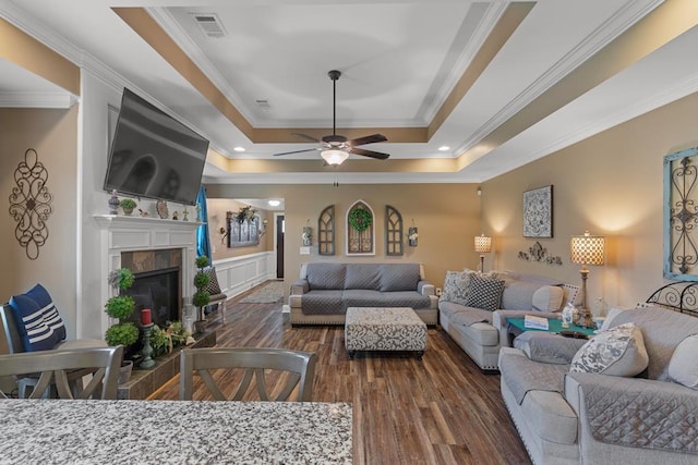 living room with ceiling fan, a raised ceiling, dark hardwood / wood-style flooring, a tiled fireplace, and ornamental molding