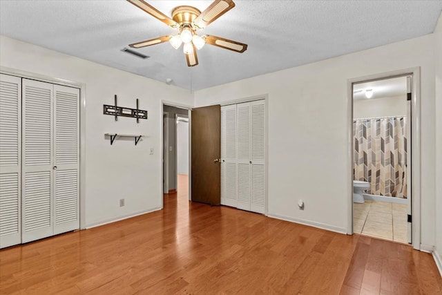 unfurnished bedroom featuring two closets, ceiling fan, light wood-type flooring, a textured ceiling, and connected bathroom