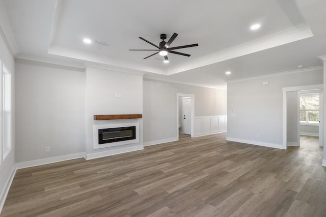 unfurnished living room with a raised ceiling, a ceiling fan, a glass covered fireplace, ornamental molding, and wood finished floors