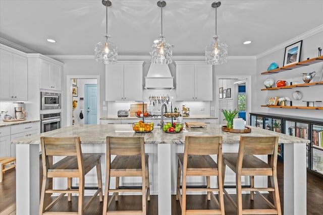 kitchen featuring stainless steel appliances, premium range hood, ornamental molding, a center island with sink, and hanging light fixtures