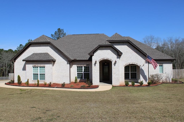 french provincial home featuring brick siding, fence, a front lawn, and roof with shingles