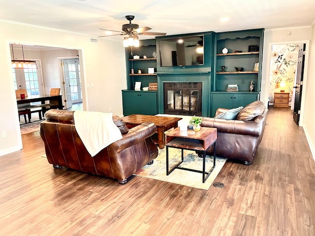 living room featuring built in shelves, ceiling fan, crown molding, and light wood-type flooring