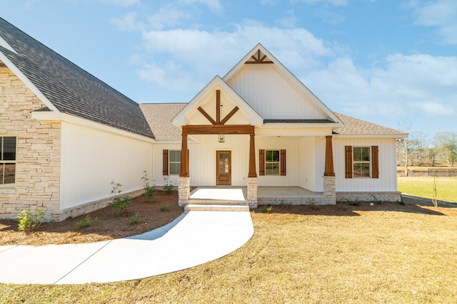 modern farmhouse with stone siding, roof with shingles, covered porch, board and batten siding, and a front yard