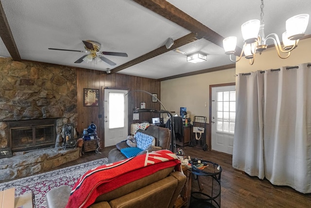 living room with wood walls, dark wood-type flooring, ceiling fan with notable chandelier, a stone fireplace, and beam ceiling