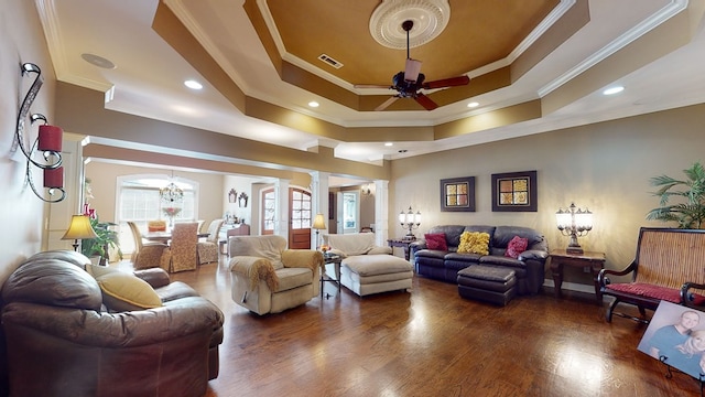 living room with dark wood-type flooring, ceiling fan with notable chandelier, ornamental molding, a tray ceiling, and decorative columns