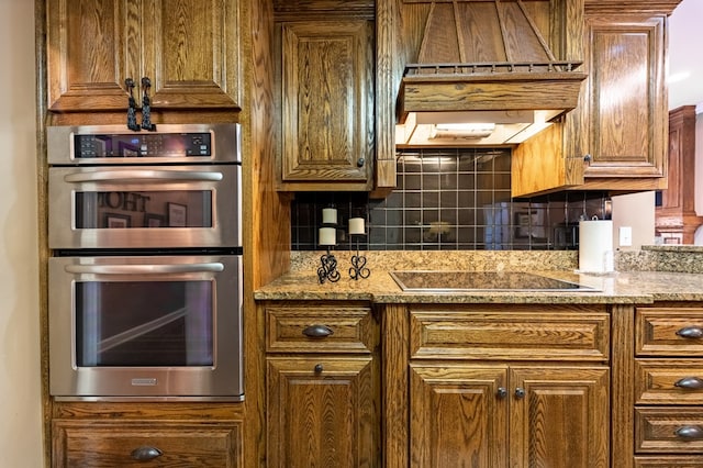 kitchen with light stone countertops, stainless steel double oven, backsplash, black electric stovetop, and exhaust hood