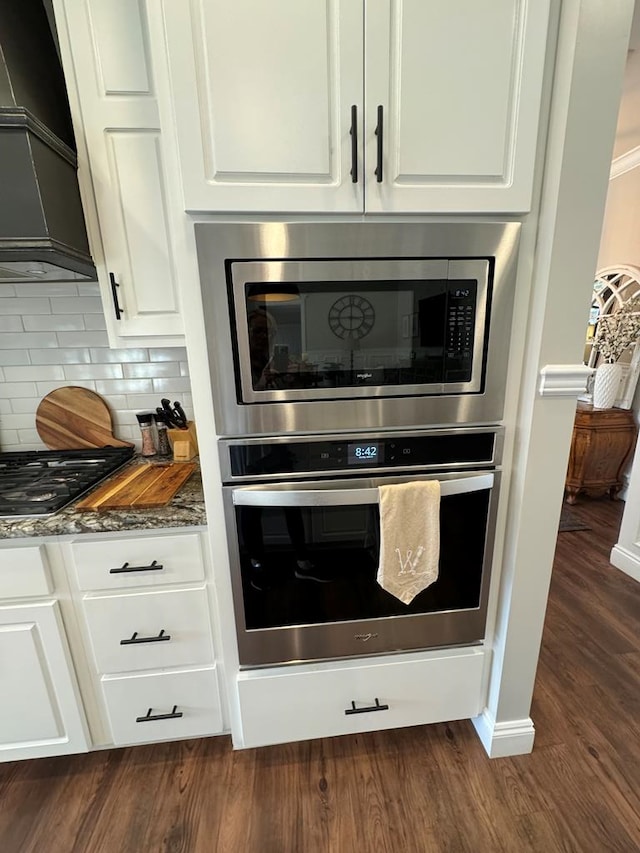 room details with tasteful backsplash, dark stone countertops, stainless steel appliances, premium range hood, and white cabinets