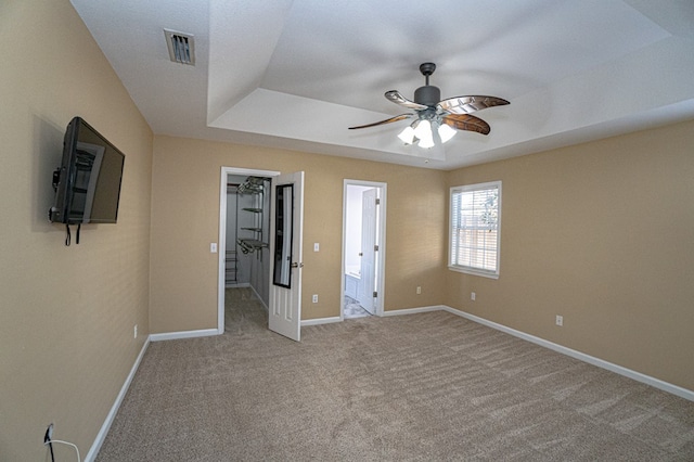 unfurnished bedroom featuring ceiling fan, light colored carpet, a raised ceiling, ensuite bath, and a spacious closet