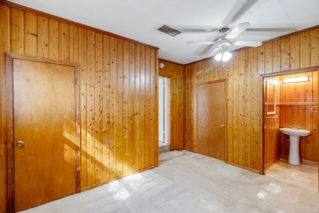 unfurnished bedroom with ensuite bath, ceiling fan, wood walls, light carpet, and ornamental molding