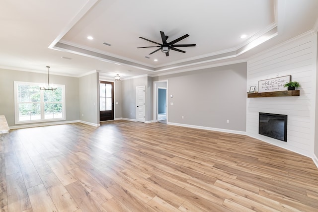 unfurnished living room featuring a large fireplace, crown molding, light hardwood / wood-style floors, a tray ceiling, and ceiling fan with notable chandelier