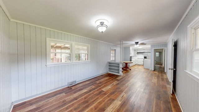 unfurnished living room with wooden walls, crown molding, ceiling fan, and dark wood-type flooring