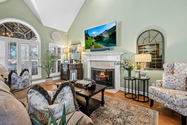 living area with high vaulted ceiling, baseboards, a tiled fireplace, and wood finished floors