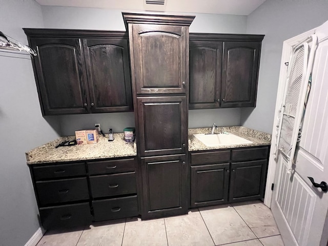 kitchen with light tile patterned flooring, light stone countertops, dark brown cabinetry, and sink