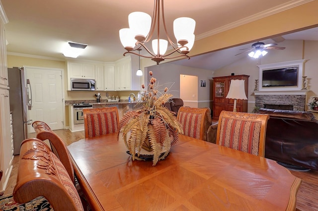dining area featuring a chandelier, light wood-style flooring, a stone fireplace, a ceiling fan, and crown molding