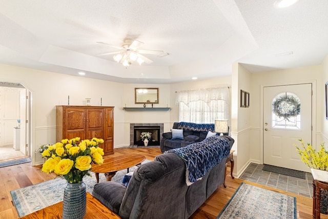 living area with arched walkways, a raised ceiling, a tiled fireplace, light wood-style flooring, and a textured ceiling