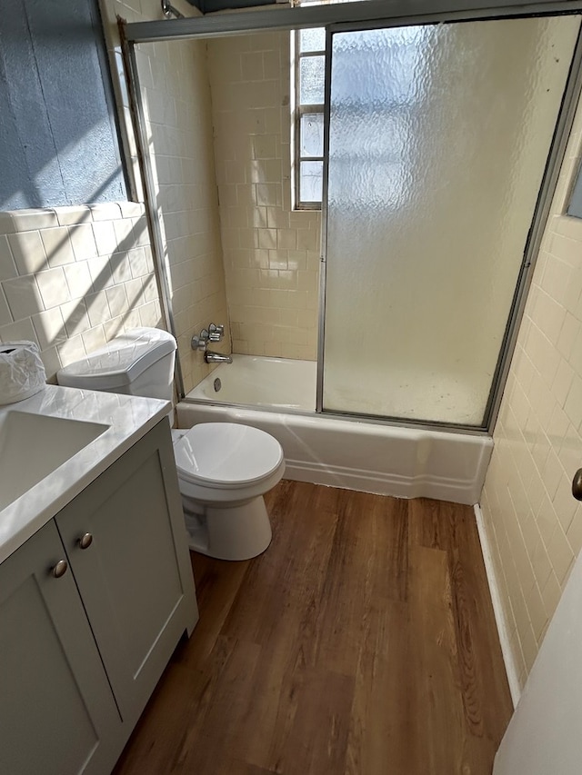 full bathroom with combined bath / shower with glass door, vanity, wood-type flooring, tile walls, and toilet