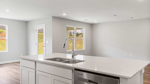 kitchen with dishwasher, a kitchen island with sink, sink, light wood-type flooring, and light stone counters