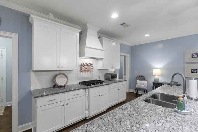 kitchen with custom exhaust hood, stainless steel gas cooktop, light stone counters, sink, and white cabinetry